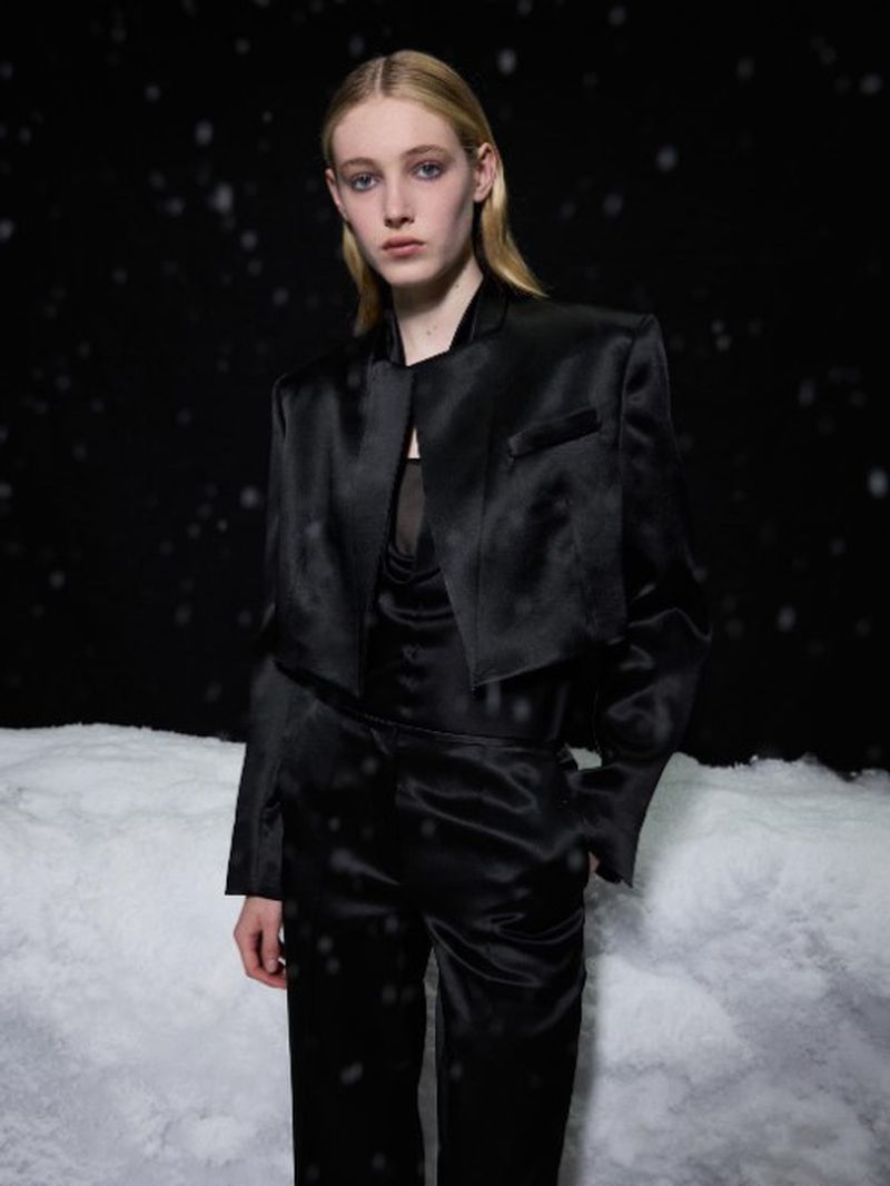 A young woman poses in a stylish, satin black pantsuit, set against a snowy, dark backdrop, showcasing a blend of fashion and winter elegance.