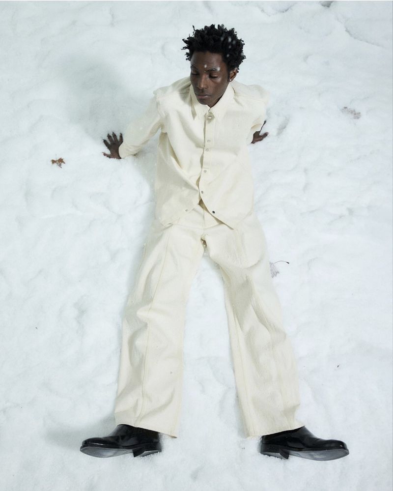 A Black adult male model in an off-white suit poses while lying on a field of puffy white material against a white background in a studio.