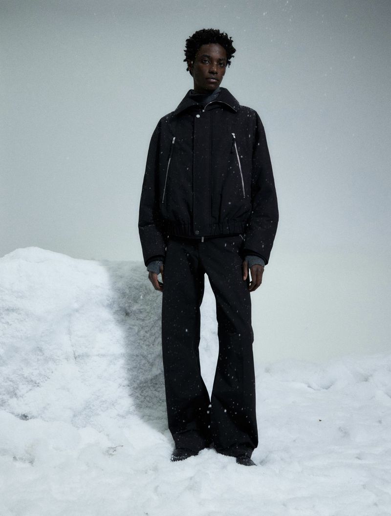 A young Black male model in a black puffer jacket and loose black pants stands in the snow in a studio. Fashion, winter clothing.