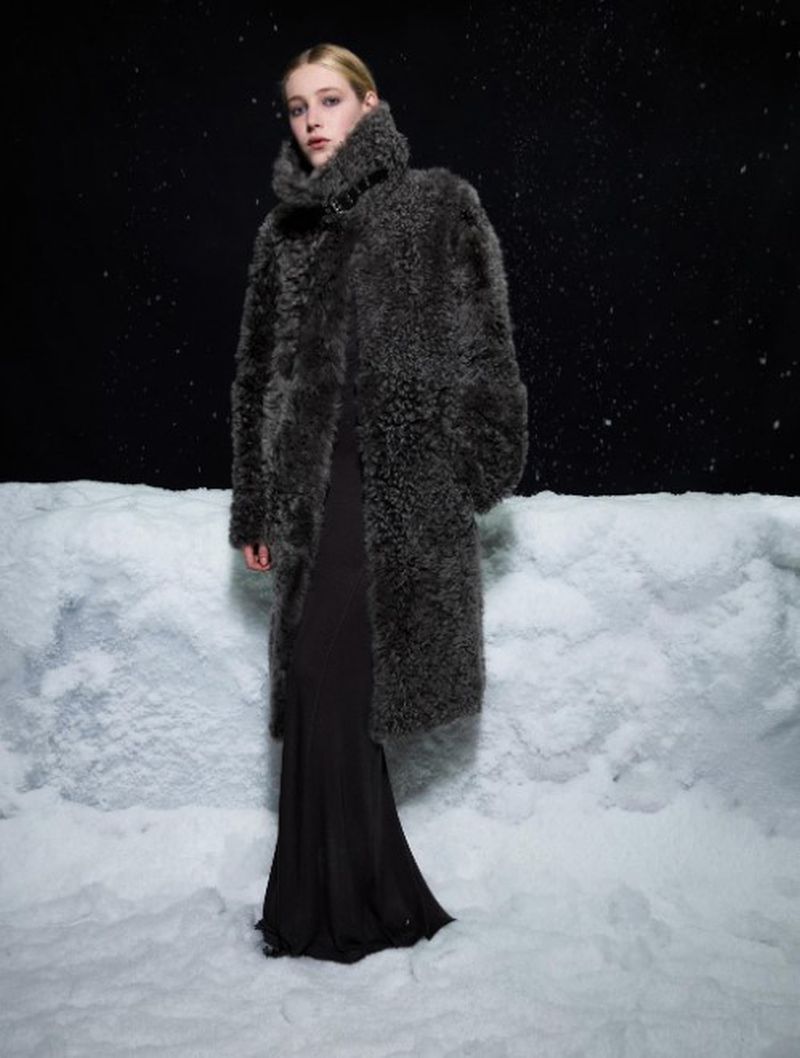 Fashionable blonde model wears a grey textured, full-length coat in front of a faux snowscape against a dark, spotted background.