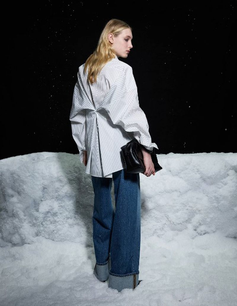 Fashion shot of a blonde adult female model wearing a white pinstripe shirt and blue jeans, holding a black clutch, against a dark background.