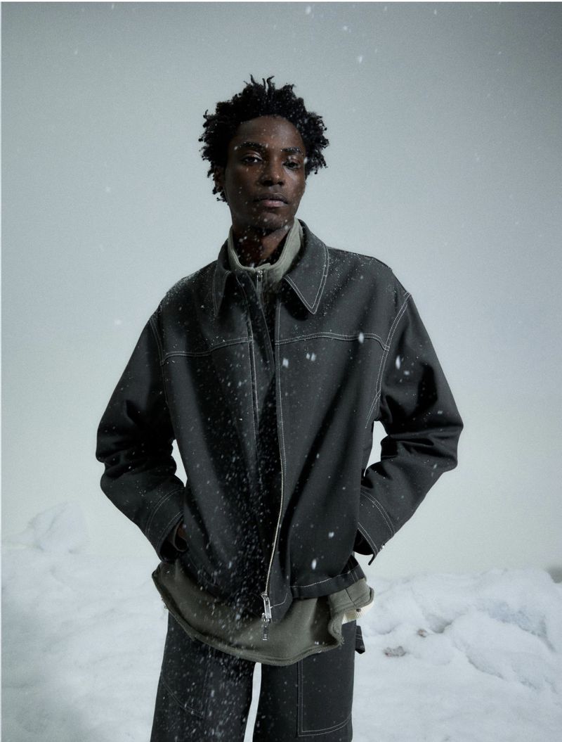 Person in a dark jacket and pants with white stitching, standing in the snow, medium shot, neutral expression, winter fashion.