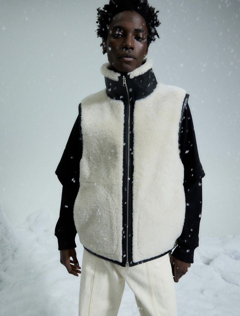 Adult male in a winter fashion shot. He is wearing a sleeveless, white, fleece jacket with a black zipper in a snowy environment.