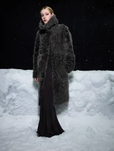 Model wearing a thick textured grey coat and long dark gown standing against a snowy backdrop at night.