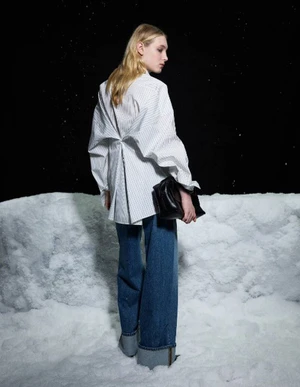 Model standing on a snowy terrain wearing an oversized striped white shirt and wide-leg blue jeans holding a black handbag.