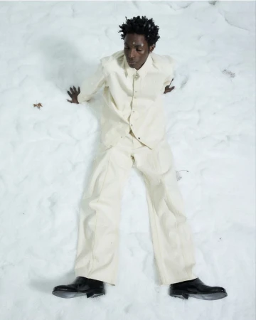 A Black male model wearing an off-white button-down shirt and matching trousers reclining against snow in black boots.