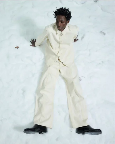 A Black male model wearing an off-white button-down shirt and matching trousers reclining against snow in black boots.