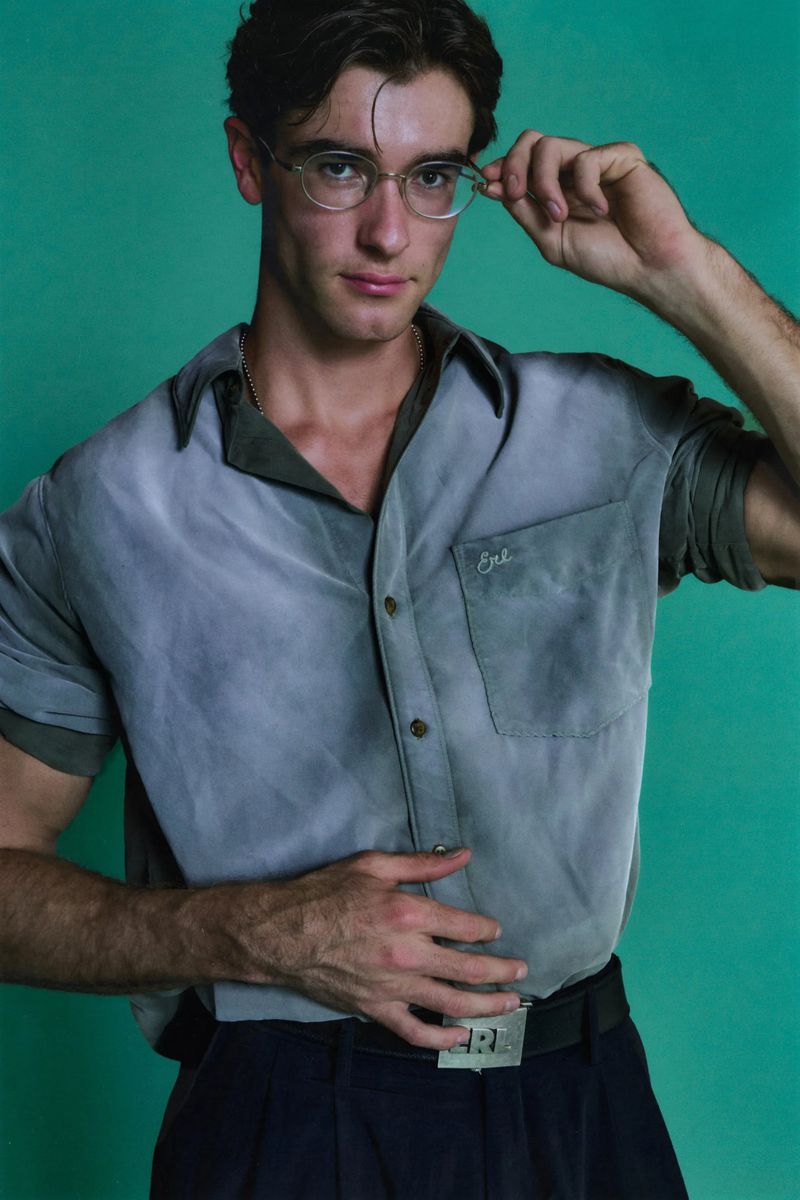 Headshot of adult male adjusting glasses, wearing grey button-down shirt and dark pants against a studio teal backdrop. Modern style.