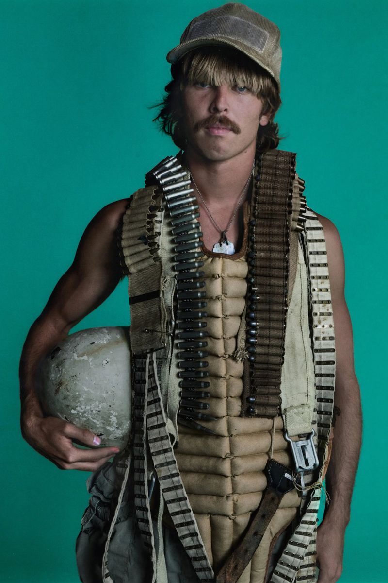 A man with a mustache and aviator cap, wearing a flight suit, is holding a helmet in a studio with a turquoise background.