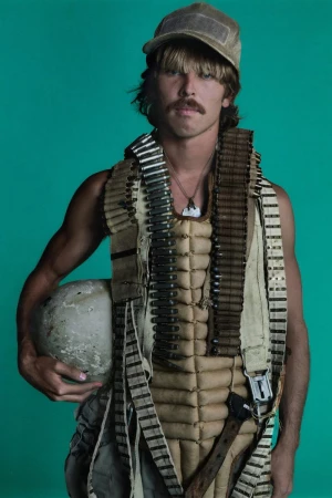 A man with a mustache and aviator cap, wearing a flight suit, is holding a helmet in a studio with a turquoise background.
