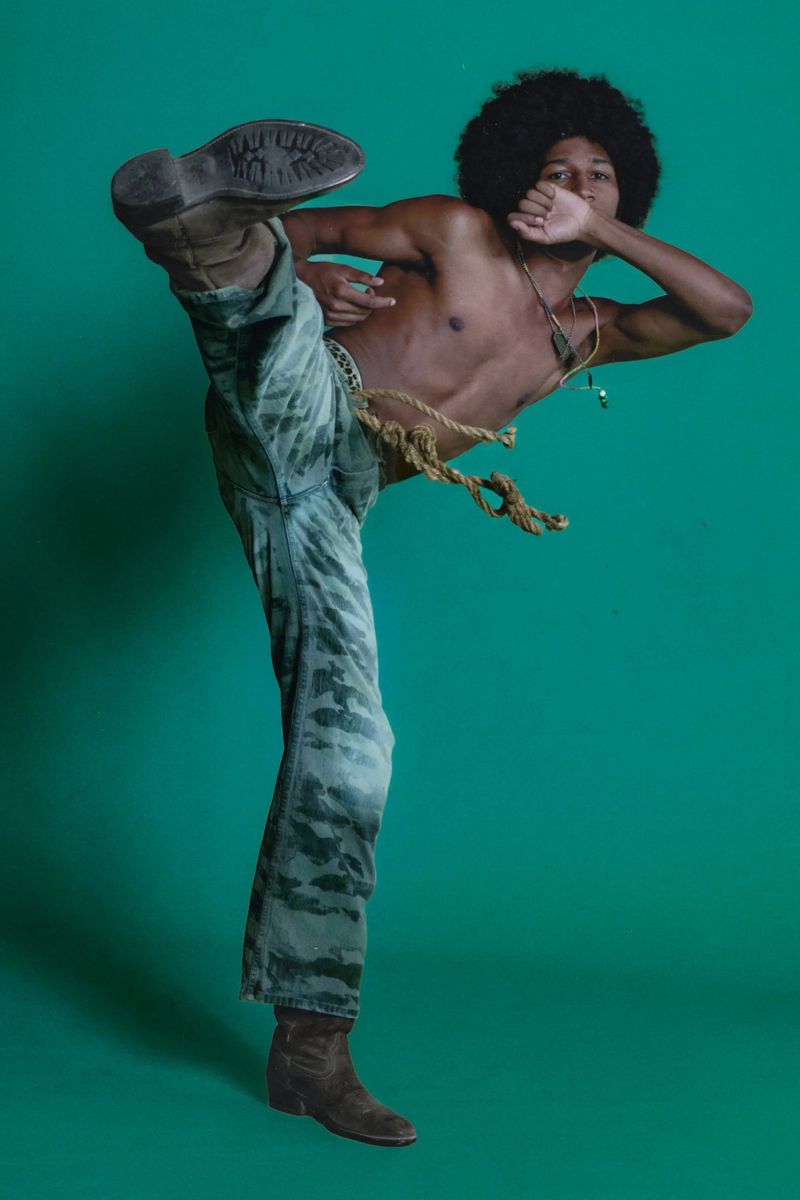 Shirtless man with an afro and patterned pants kicks in a photo studio setting with a green backdrop, captured in a medium shot.