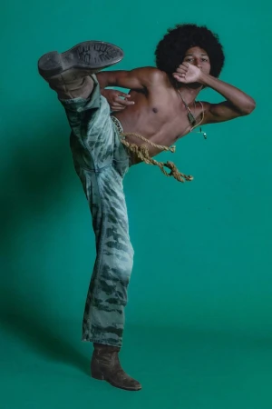 Shirtless man with an afro and patterned pants kicks in a photo studio setting with a green backdrop, captured in a medium shot.