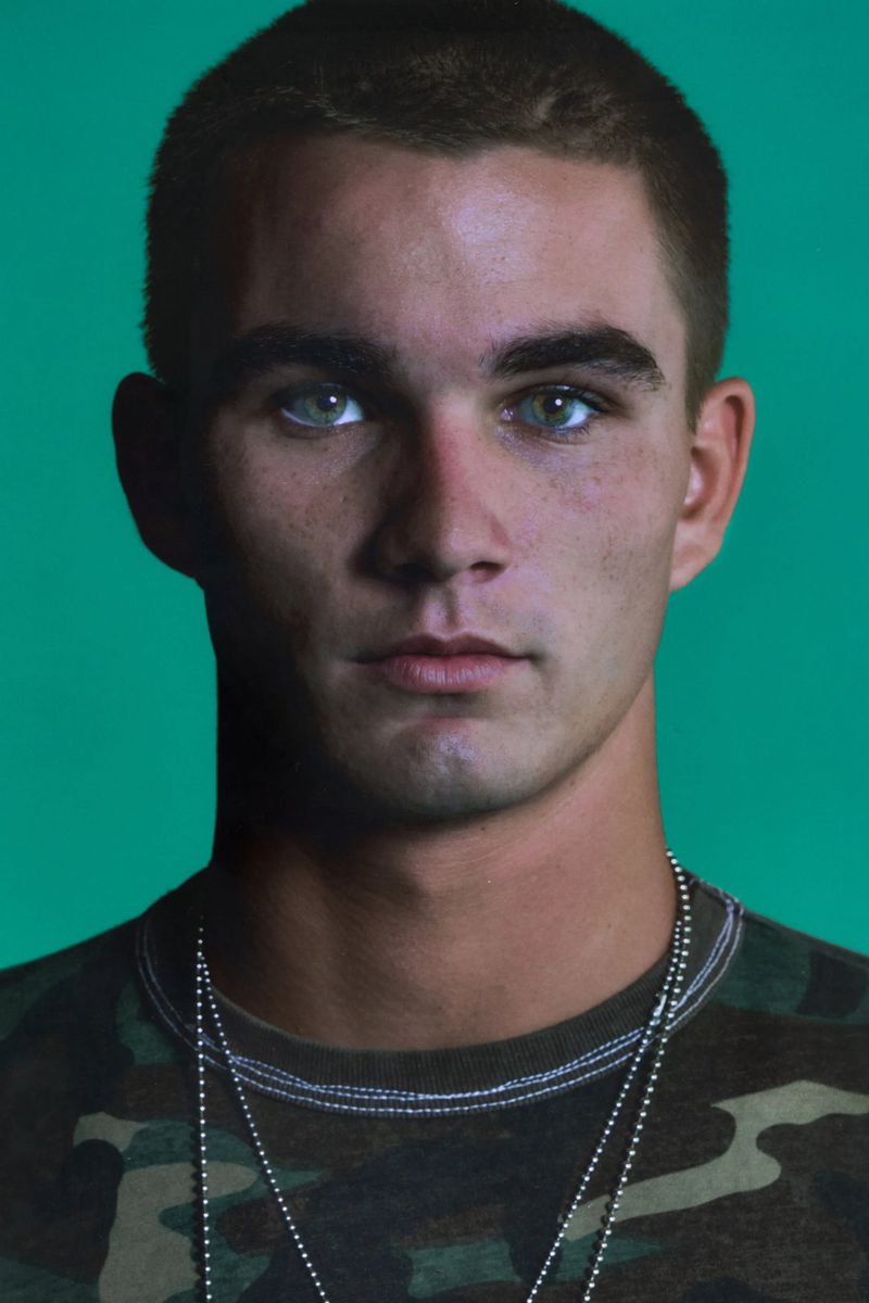 Close-up portrait of a young adult male with fair skin and short brown hair wearing a camouflage t-shirt against a solid green backdrop.