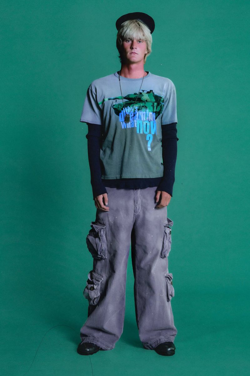 Full-length shot of a young adult male with blond hair wearing a graphic t-shirt, wide leg cargo pants, and a small hat against green backdrop.