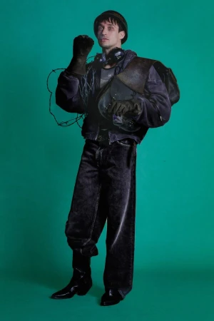 Man wearing a purple jacket, dark denim jeans, hat, and gloves against a solid green background for ERL Resort Spring Summer 2026.