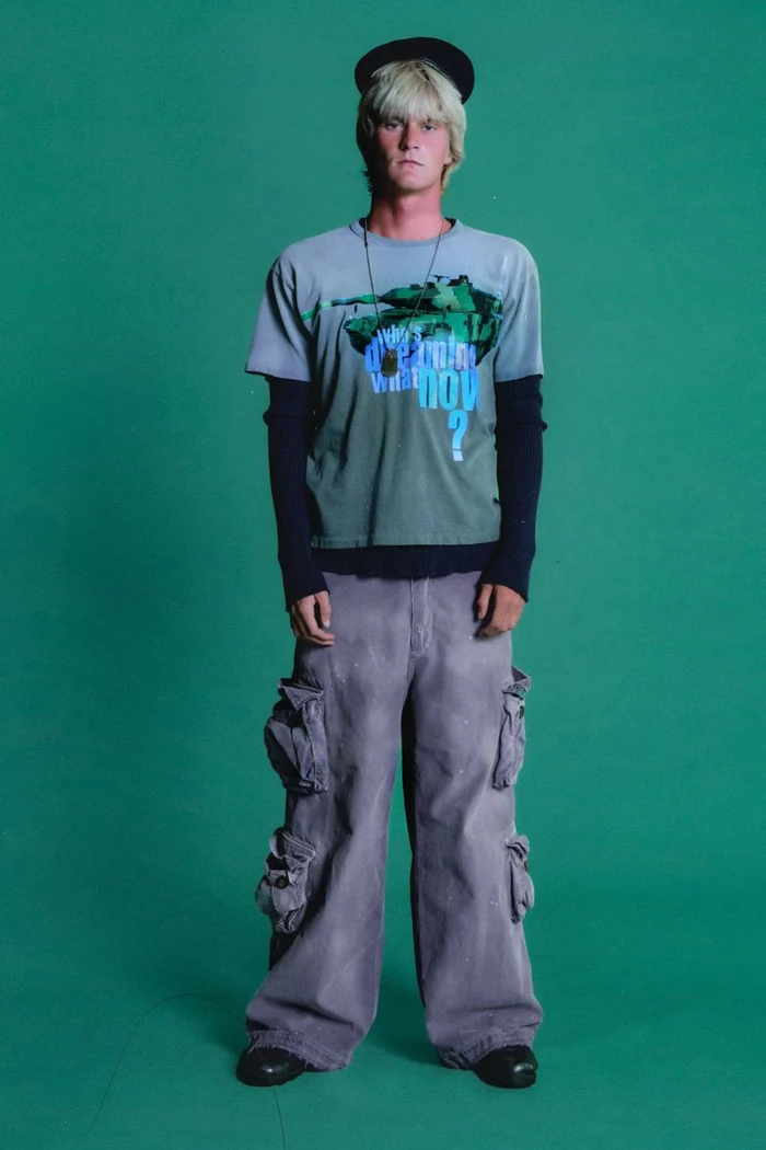 Model wearing a graphic t-shirt over long sleeves and baggy cargo pants against a green seamless studio backdrop.