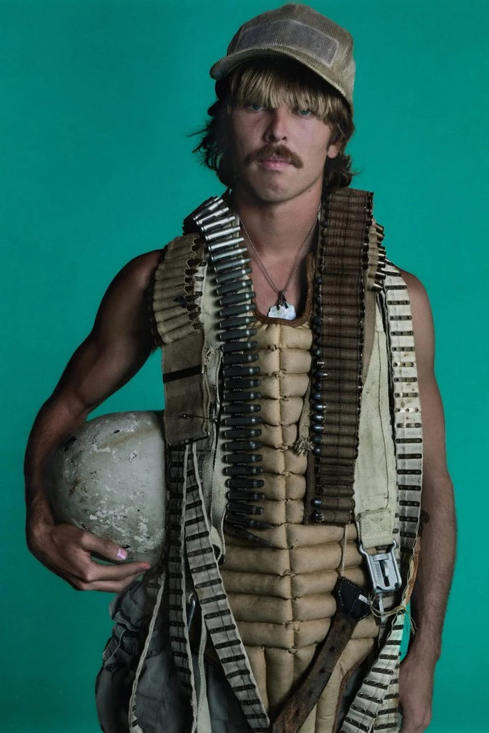 Man wearing a padded vest with bandoliers and a cap holding a helmet against a solid teal background.