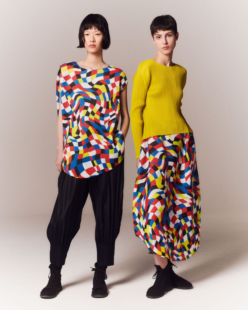 Two models pose in a photo studio wearing colorful, geometric print tops and bottoms and black boots against a pale backdrop.