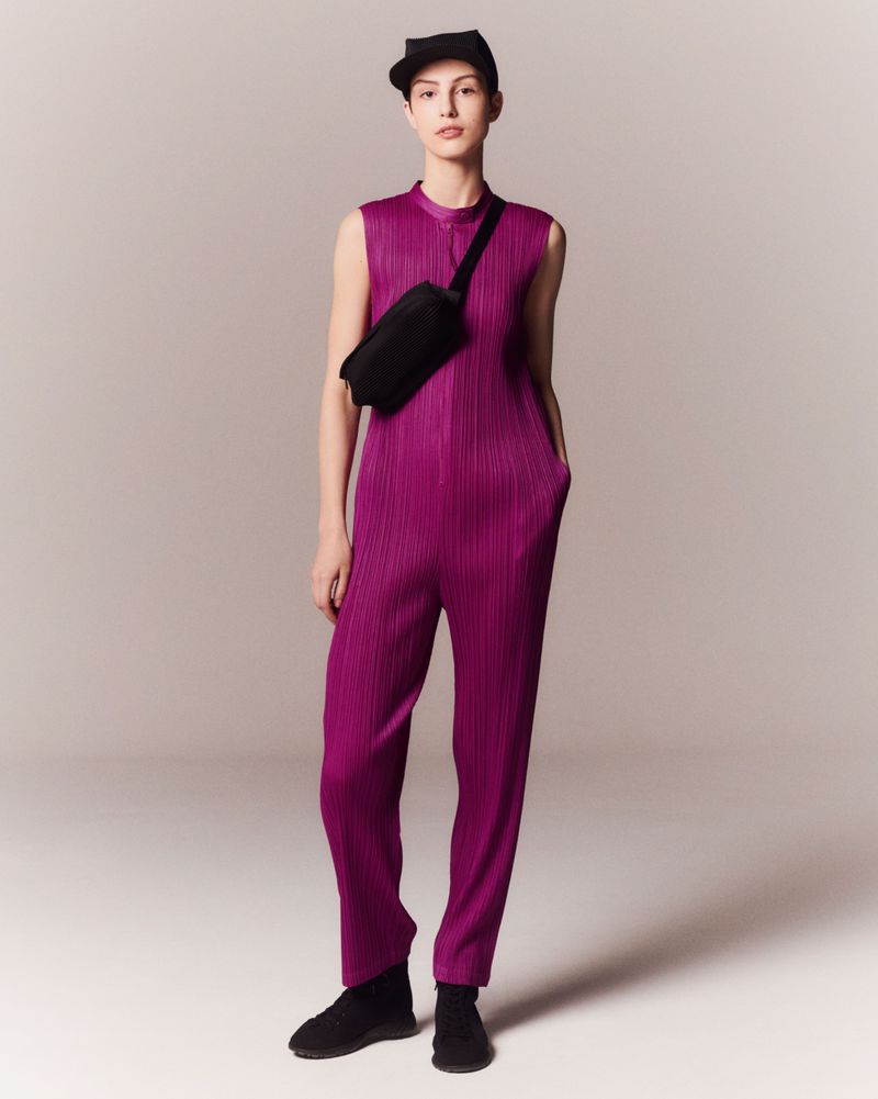 Fashion model posing in a purple pleated jumpsuit with a black fanny pack and cap, standing in a neutral studio setting with soft lighting.