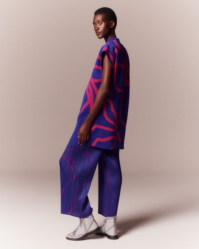 An adult female model is posing in a photo studio wearing a blue and pink printed top and pants with white boots in a studio shot.