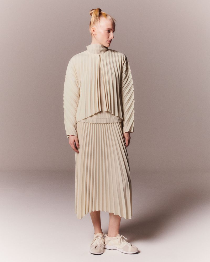 A slender woman stands in a neutral studio, wearing a pleated top and skirt with matching sneakers, and her hair in a topknot.