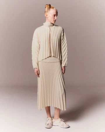 A slender woman stands in a neutral studio, wearing a pleated top and skirt with matching sneakers, and her hair in a topknot.