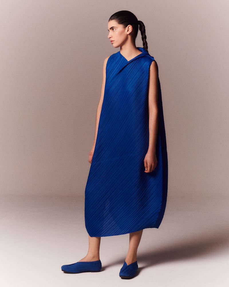 Full-length shot of a woman posing in a pleated blue dress and matching flats, standing in front of a gradient cream-colored backdrop.