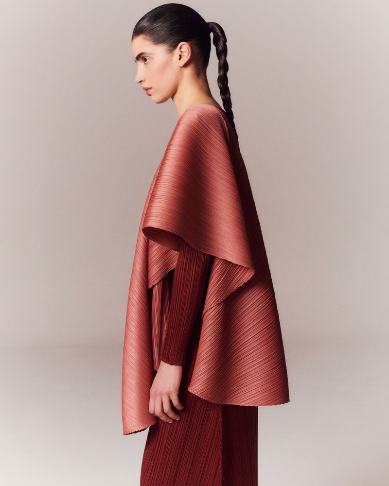 A woman in a pleated, earth-tone outfit stands in profile against a neutral backdrop, showcasing the cape-like design and fabric texture.