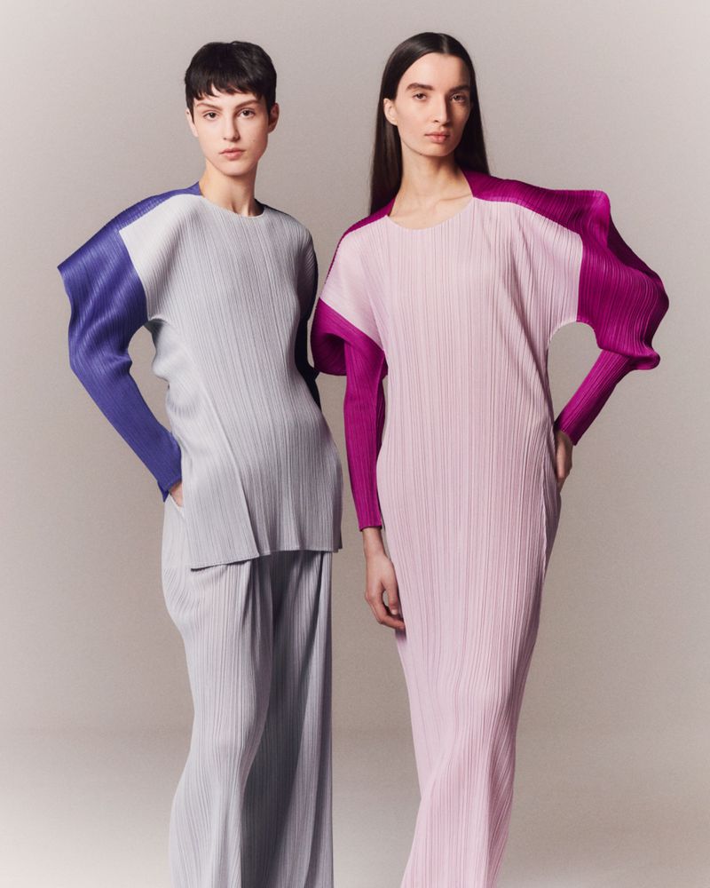 Two fashion models pose in a studio wearing pleated long sleeve tops, loose fitting pants, and a dress in shades of purple and grey.