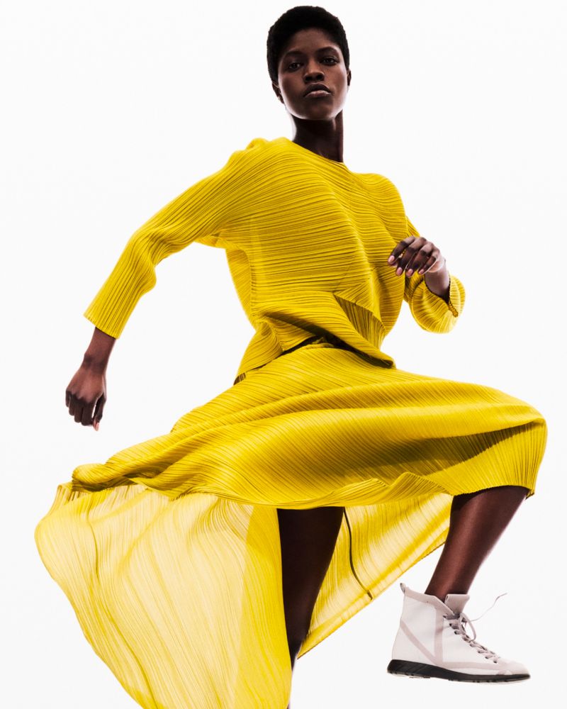 Fashion shot of Black female model in yellow pleated matching top and skirt with white sneakers, posing on white studio backdrop.