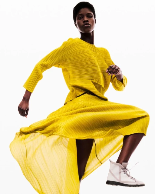 Fashion shot of Black female model in yellow pleated matching top and skirt with white sneakers, posing on white studio backdrop.