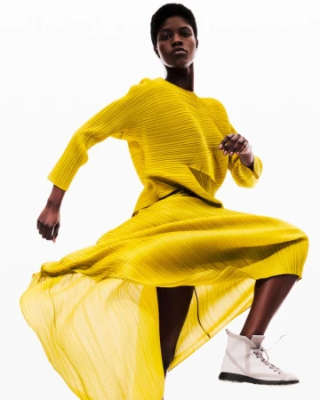 Fashion shot of Black female model in yellow pleated matching top and skirt with white sneakers, posing on white studio backdrop.