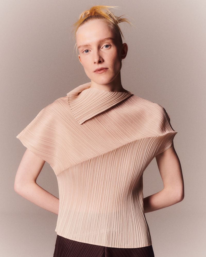 Blonde woman posing in a pleated top against a neutral backdrop. Fashion shot features a textured garment and a minimalist aesthetic.