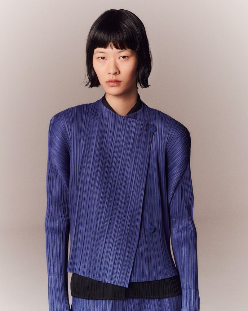 Asian woman in a vertically-pleated, two-button, wrap-style blue jacket against an off-white backdrop. Fashion editorial, medium shot.