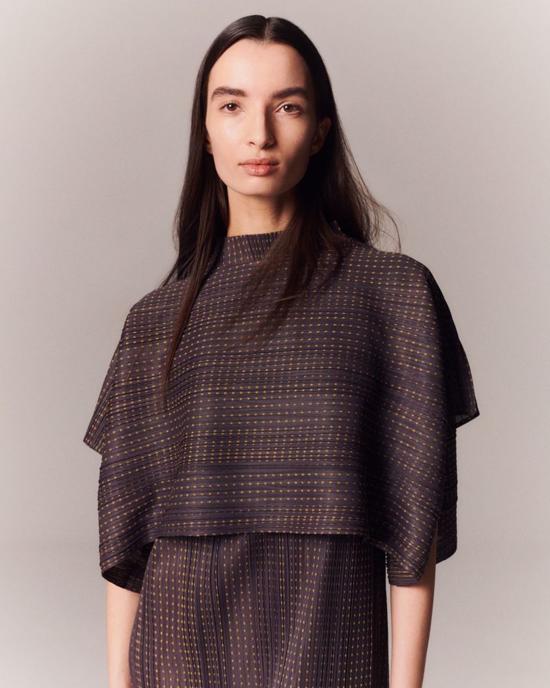 A woman with long dark hair poses indoors in a textured brown top and skirt. The portrait has a soft, neutral color palette.