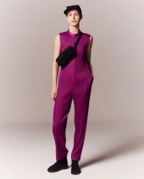 Model wearing a magenta pleated jumpsuit with a black fanny pack and cap standing against a pale grey seamless background