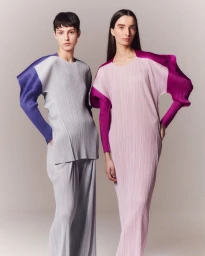 Two female models wearing pleated Issey Miyake garments against a light grey seamless studio background.