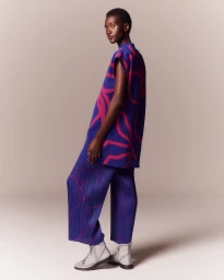 Model wearing a blue patterned tunic and pleated trousers with grey boots against a neutral backdrop.
