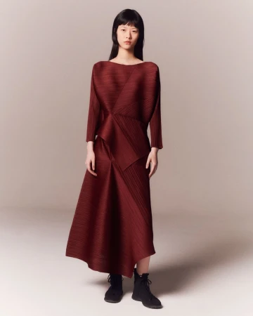 Model wearing a pleated burgundy long sleeve dress and black ankle boots against a neutral grey studio background