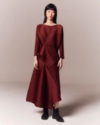 Model wearing a pleated burgundy long sleeve dress and black ankle boots against a neutral grey studio background