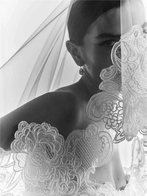 Monochrome portrait of a bride with fair skin, partially obscured by an ornate, lacy veil, adorned with an earring on display.