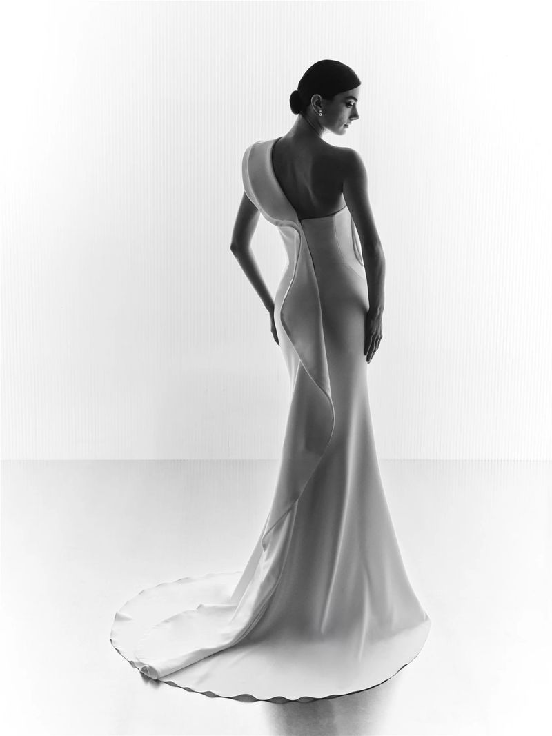 Elegant woman in white one-shoulder gown, stands in a brightly lit studio. Formal attire, minimalist backdrop, classic updo hairstyle.