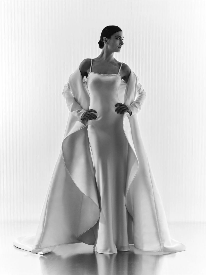 Elegant adult female in a satin slip dress and jacket, hands on hips, posing against a softly lit, vertical-striped background in a studio setting.