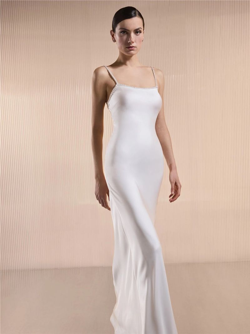 Elegant woman in a full-length white dress with spaghetti straps stands against a light background in a fashion studio shot.
