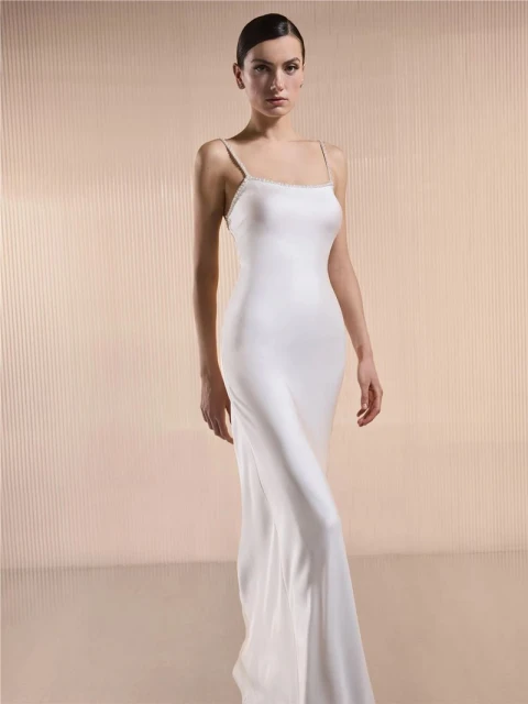Elegant woman in a full-length white dress with spaghetti straps stands against a light background in a fashion studio shot.
