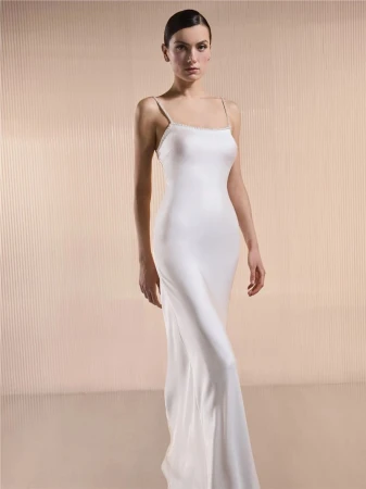 Elegant woman in a full-length white dress with spaghetti straps stands against a light background in a fashion studio shot.