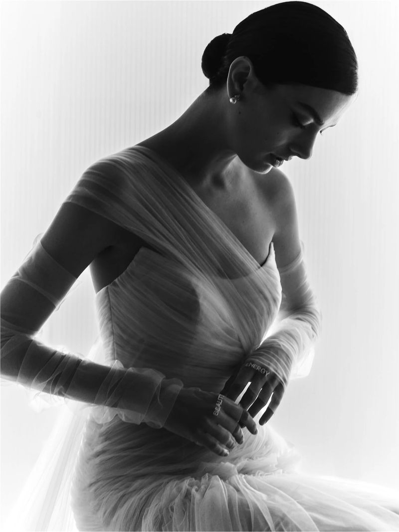 Monochrome portrait of a woman with dark hair in an updo, wearing a light-colored, draped gown with sheer sleeves, against a bright background.