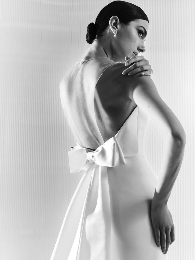 Elegant backless gown with a large bow detail, worn by a woman with an updo in a studio setting, captured in a monochrome shot.