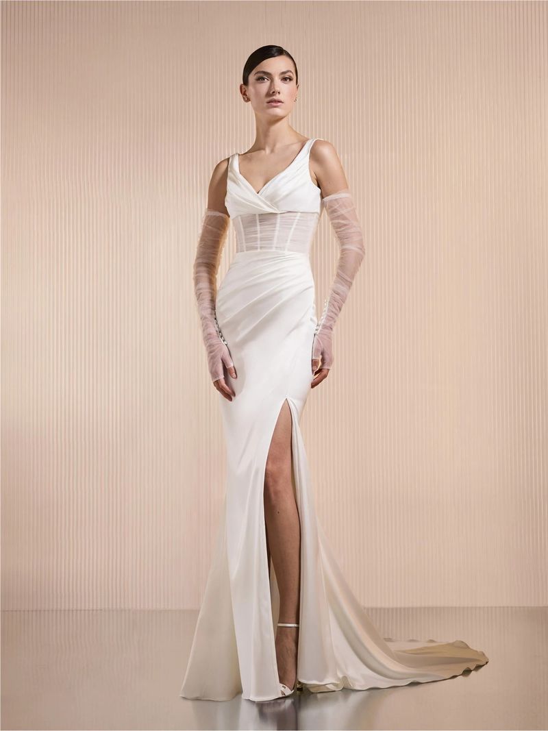 Elegant woman in white V-neck slit gown with sheer sleeves poses in studio, radiating sophistication and timeless bridal fashion.