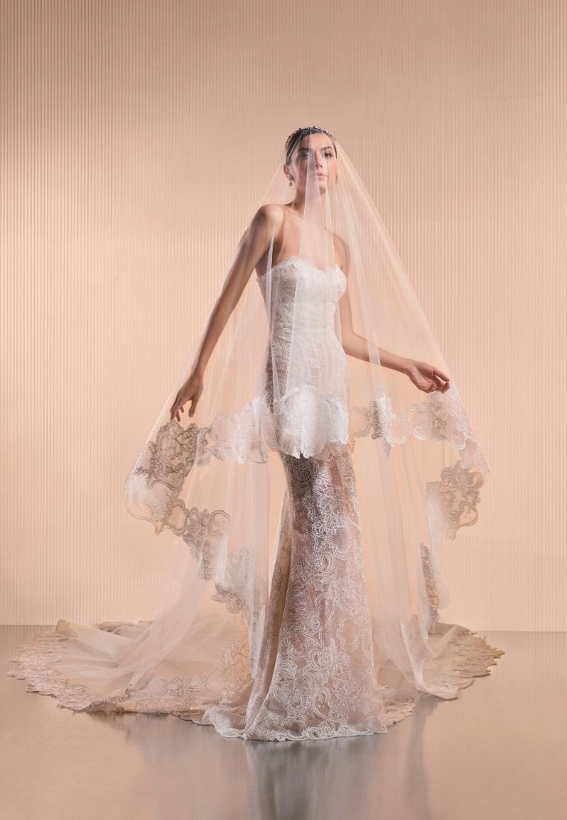 Woman in an ornate white lace wedding gown and veil poses in a studio, exuding elegance and sophistication in a full shot.